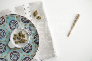 Top Down View on Marijuana Buds on a Vintage Plate and Joint atop a Silver Placemat and White Background Minimalist Cannabis - The Cannabiz Agency