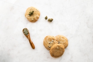 Top Down View on White Marble of Edible Marijuana Chocolate Chip Cookies, Cannabis Buds and Ground Weed on a Wooden Spoon - The Cannabiz Agency